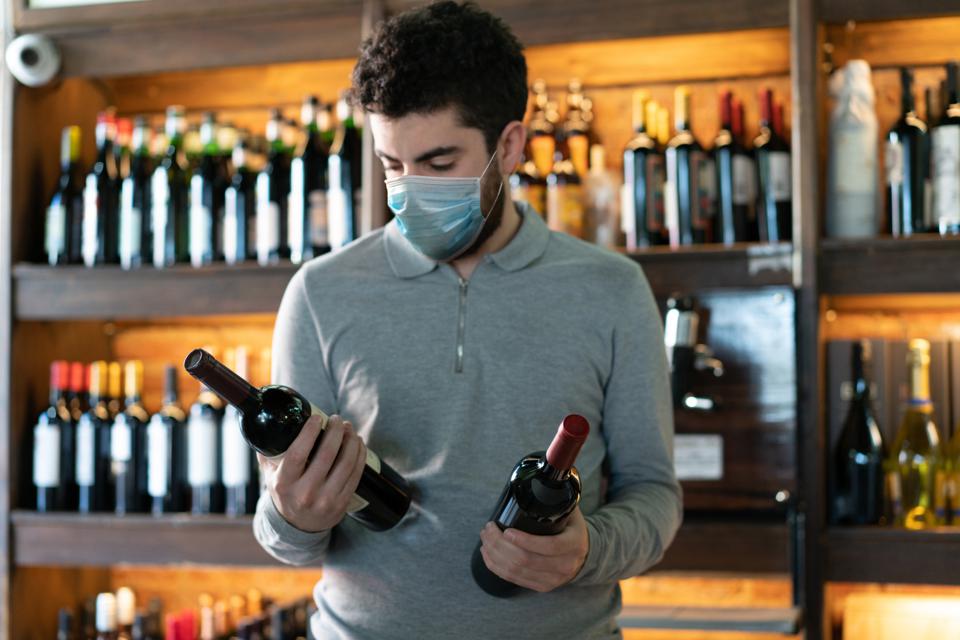 Young man at a wine cellar choosing between two wine bottles wearing a protective face mask